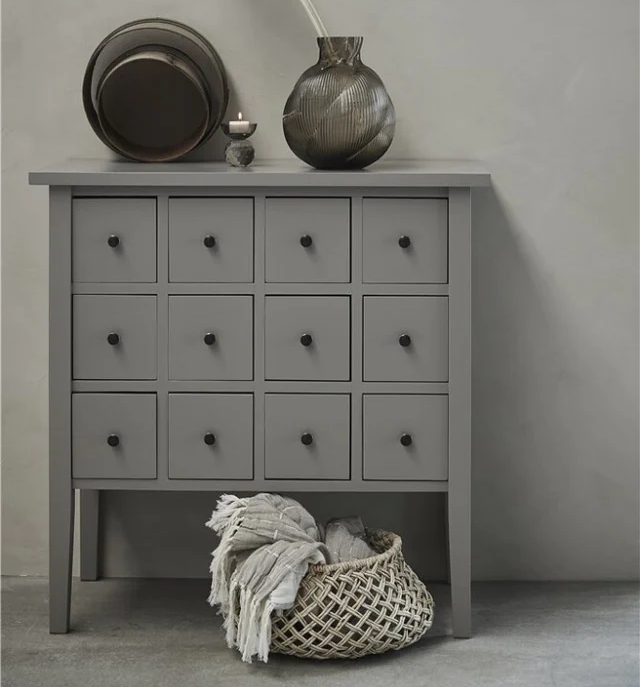 Grauer Schrank mit Schubladen, Vase und Trockenblume auf einer neutralen Wand.